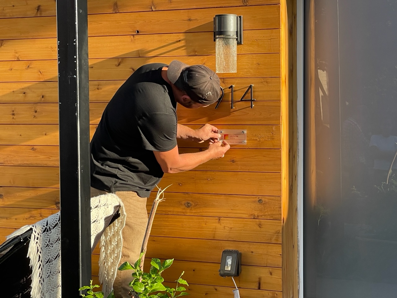 A builder mounting a Passive House certification plaque on a wood-clad wall.